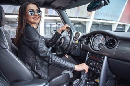 Photo for Beautiful girl in leather jacket and sun glasses is smiling while driving a car - Royalty Free Image