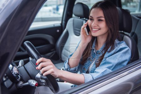 Foto de Hermosa chica en chaqueta de jean está hablando en el teléfono móvil y sonriendo mientras está sentado en un coche - Imagen libre de derechos