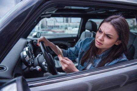 Photo for Beautiful girl in jean jacket is using a smart phone and looking forward with unsatisfied look while driving a car - Royalty Free Image
