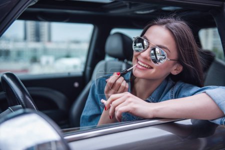 Téléchargez les photos : Belle fille en jean veste et lunettes de soleil applique brillant à lèvres et souriant tout en étant assis dans sa voiture - en image libre de droit