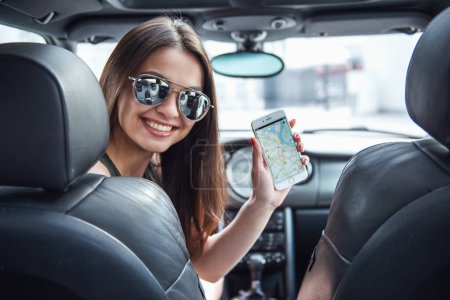 Téléchargez les photos : Belle fille en lunettes de soleil montre un téléphone intelligent avec carte numérique, regardant l'appareil photo et souriant tout en étant assis dans sa voiture - en image libre de droit