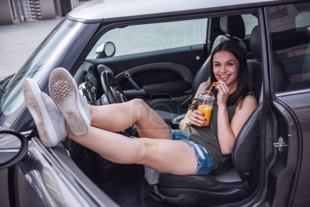 Photo for Beautiful girl is drinking juice, looking at camera and smiling while sitting in her car - Royalty Free Image