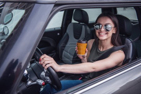 Photo for Beautiful girl in sun glasses is drinking coffee, looking at camera and smiling while sitting in her car - Royalty Free Image