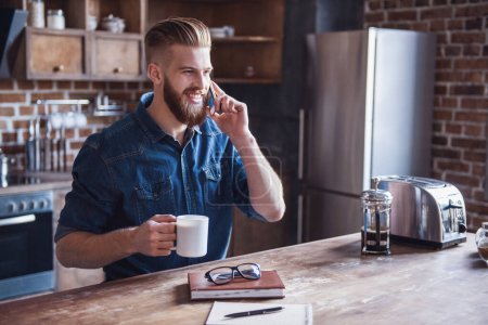 Téléchargez les photos : Beau jeune homme barbu parlant sur le téléphone portable, buvant du café et souriant tout en se reposant dans la cuisine - en image libre de droit