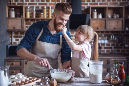 Photo for Cute little girl and her handsome bearded dad in aprons are preparing dough and smiling while cooking in kitchen - Royalty Free Image