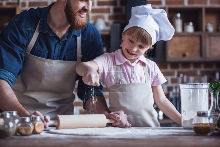 Téléchargez les photos : Petite fille mignonne et son beau papa barbu dans des tabliers aplatissent la pâte à l'aide d'un rouleau à pâtisserie et souriant tout en cuisinant dans la cuisine - en image libre de droit