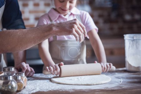 Téléchargez les photos : Image recadrée de la petite fille et de son beau père dans des tabliers aplatissant la pâte à l'aide d'un rouleau à pâtisserie tout en cuisinant dans la cuisine - en image libre de droit