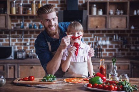 Photo for Cute little girl and her handsome bearded dad in aprons are having fun with ingredients while cooking pizza in kitchen - Royalty Free Image