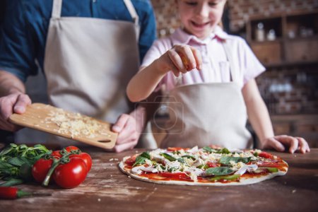 Téléchargez les photos : Petite fille mignonne et son beau papa dans des tabliers sourient tout en cuisinant des pizzas dans la cuisine - en image libre de droit