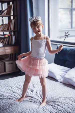 Foto de Linda niña en falda de tul, con corona y varita mágica está mirando a la cámara y sonriendo mientras juega en su habitación - Imagen libre de derechos
