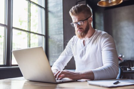 Téléchargez les photos : Jeune homme barbu dans des lunettes en utilisant un ordinateur portable tout en travaillant dans le bureau - en image libre de droit