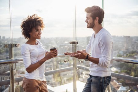 Foto de Hermosa pareja joven está hablando, tomando café y sonriendo mientras está de pie en el balcón del edificio de la ciudad - Imagen libre de derechos