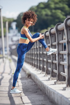 Photo for Beautiful girl in sportswear and earphones is looking at camera and smiling while stretching before morning run - Royalty Free Image