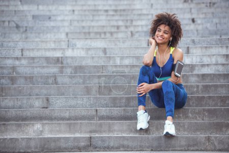 Téléchargez les photos : Belle fille en vêtements de sport et écouteurs regarde loin et sourit tout en se reposant sur les escaliers pendant la course du matin - en image libre de droit