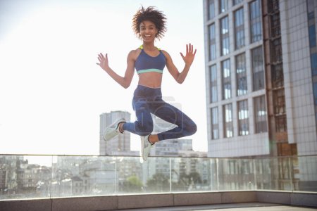 Photo for Beautiful girl in sportswear is looking at camera and smiling while jumping on city background - Royalty Free Image