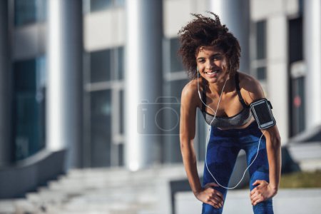 Foto de Hermosa chica en ropa deportiva y auriculares está mirando a la cámara y sonriendo mientras se apoya en sus rodillas durante la carrera de la mañana - Imagen libre de derechos