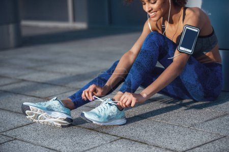 Photo for Beautiful girl in sportswear and earphones is lacing running shoes and smiling during morning run - Royalty Free Image