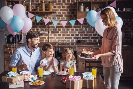 Foto de Familia feliz está sentado en la mesa en la cocina decorada durante la celebración del cumpleaños, mamá está sosteniendo un pastel de cumpleaños - Imagen libre de derechos
