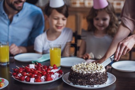 Foto de La familia feliz está sentada a la mesa en la cocina decorada durante la celebración del cumpleaños. Imagen recortada de mamá cortando pastel de cumpleaños - Imagen libre de derechos