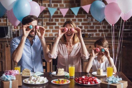 Photo for Family celebrating birthday in decorated kitchen. Mom, dad and son are holding macarons and smiling - Royalty Free Image