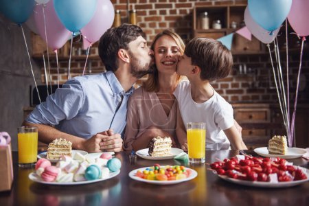 Foto de Familia celebrando el cumpleaños de la madre en la cocina decorada. Hermosa mujer está mirando a la cámara y sonriendo mientras marido e hijo la besan en las mejillas - Imagen libre de derechos