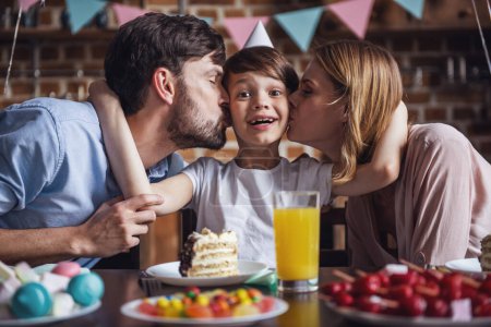 Téléchargez les photos : Famille célébrant l'anniversaire du fils dans la cuisine décorée. Mignon petit garçon regarde la caméra et souriant pendant que maman et papa l'embrassent dans les joues - en image libre de droit