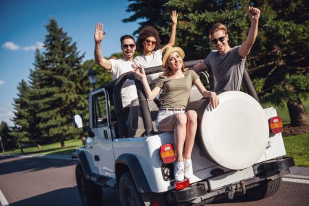 Téléchargez les photos : Les jeunes gens heureux saluent la caméra et sourient en voyageant en voiture par temps ensoleillé - en image libre de droit