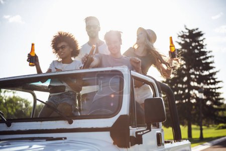 Téléchargez les photos : Les jeunes gens heureux tiennent des bouteilles de boissons et sourient en voyageant en voiture par temps ensoleillé - en image libre de droit