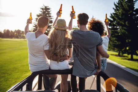 Téléchargez les photos : Vue arrière de jeunes gens heureux tenant des bouteilles de boisson et parlant en voiture par temps ensoleillé - en image libre de droit
