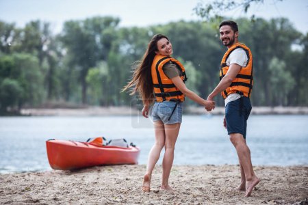 Photo for Happy young couple in sea vests are holding hands, looking at camera and smiling after sailing a kayak - Royalty Free Image