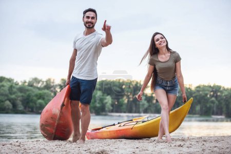 Foto de Feliz joven pareja está hablando y sonriendo mientras tirando de kayaks en la playa después de navegar - Imagen libre de derechos