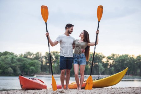 Photo for Happy young couple is holding paddles and smiling while standing near the kayaks on the beach after sailing - Royalty Free Image