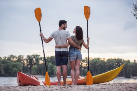 Téléchargez les photos : Vue de l'arrière du jeune couple tenant des pagaies et s'embrassant debout près des kayaks sur la plage et regardant le coucher du soleil - en image libre de droit