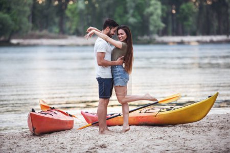 Foto de Feliz joven pareja está abrazando y sonriendo mientras está de pie cerca de los kayaks en la playa después de navegar - Imagen libre de derechos