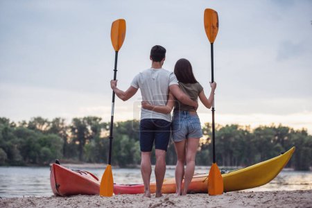 Foto de Vista trasera de la joven pareja sosteniendo paletas y abrazándose mientras está de pie cerca de los kayaks en la playa y viendo la puesta de sol - Imagen libre de derechos