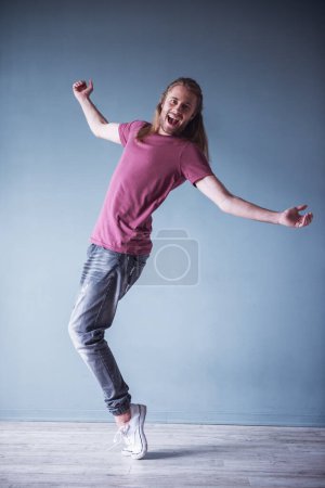 Téléchargez les photos : Jeune homme élégant avec les cheveux blonds épaule longueur et dans des vêtements décontractés pose à la caméra tout en se tenant sur les orteils, sur fond gris - en image libre de droit