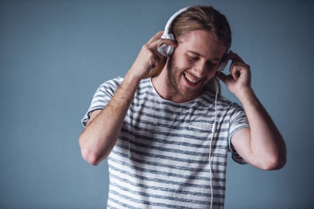 Foto de Joven con estilo con el pelo rubio de longitud de los hombros y en los auriculares está escuchando música y sonriendo, sobre fondo gris - Imagen libre de derechos