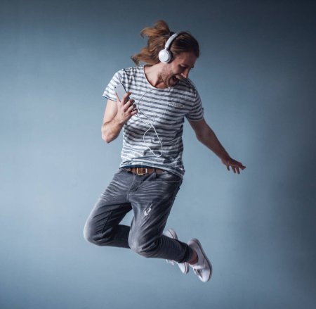 Photo for Stylish young man with shoulder-length blond hair and in headphones is listening to music while jumping, on gray background - Royalty Free Image