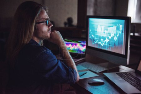 Photo for Handsome young businessman with shoulder-length blond hair and in casual wear and eyeglasses is studying graphs while working with a computer at night - Royalty Free Image