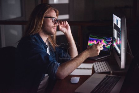 Photo for Handsome young businessman with shoulder-length blond hair and in casual wear and eyeglasses is working with a computer at night - Royalty Free Image