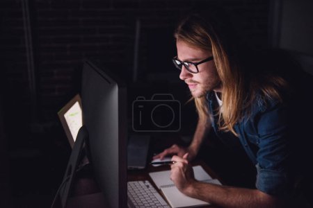 Photo for Handsome young businessman with shoulder-length blond hair and in casual wear and eyeglasses is making notes while working with a computer at night - Royalty Free Image