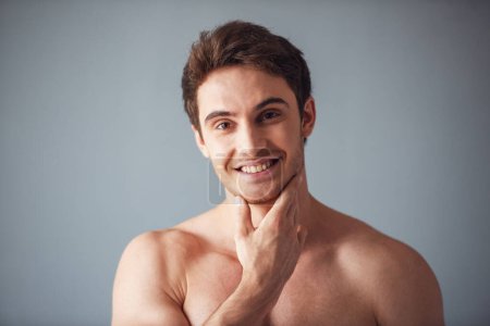 Photo for Portrait of handsome young man with bare torso touching his chin,  looking at camera and smiling, on gray background - Royalty Free Image