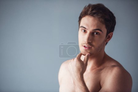 Foto de Guapo joven con el torso desnudo está tocando su barbilla y mirando seductora a la cámara, sobre fondo gris - Imagen libre de derechos