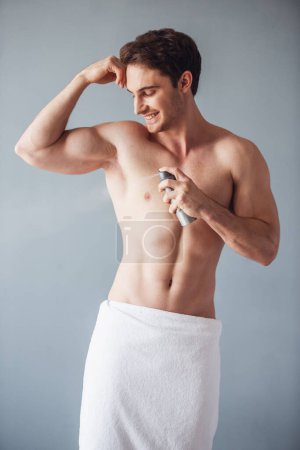 Téléchargez les photos : Beau jeune homme enveloppé dans une serviette de bain et avec torse nu utilise un déodorant spray et souriant, sur fond gris - en image libre de droit