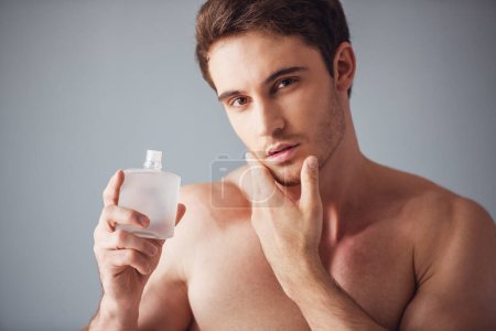 Téléchargez les photos : Beau jeune homme avec torse nu tient lotion après-rasage, toucher son menton et regarder séduisante caméra, sur fond gris - en image libre de droit