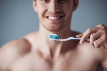 Téléchargez les photos : Image recadrée d'un beau jeune homme au torse nu tenant une brosse à dents et souriant, sur fond gris - en image libre de droit