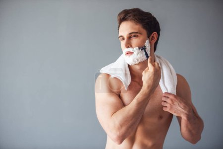 Foto de Joven guapo con espuma en la cara se afeita con una navaja de afeitar, sobre fondo gris - Imagen libre de derechos