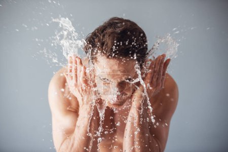 Foto de Un joven guapo con el torso desnudo se lava la cara, salpicaduras de agua alrededor, fondo gris - Imagen libre de derechos