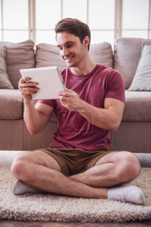 Téléchargez les photos : Beau gars en vêtements décontractés utilise une tablette numérique et souriant tout en étant assis sur le sol à la maison - en image libre de droit