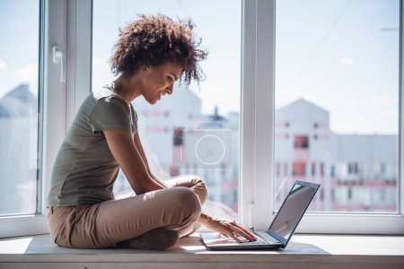 Téléchargez les photos : Belle fille travaille avec un ordinateur portable et souriant tout en étant assis sur le rebord de la fenêtre à la maison - en image libre de droit